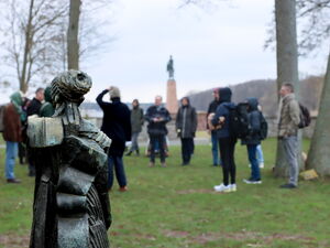 Im Vordergrund ein Denkmal. Dahinter draußen auf einer Wiese zwischen Bäumen stehen einige Menschen in einem Kreis.