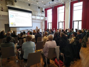 Veranstaltungssaal bei einer Konferenz oder Tagung. Teilnehmende sitzen in Reihen und vorn läuft eine Präsentation auf einer Leinwand.