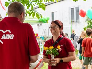 Eine Frau mit einer kleinen weißen Vase mit Blumen in den Händen, lächelt breit. Ihr gegenüber ein Mann im roten AWO Poloshirt von hinten. Kinder mit Luftballons gehen vorbei.